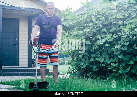 Mann in Brille, Shorts und T-Shirt mäht Rasen mit Benzin Trimmer vor dem Hintergrund des Weinbergs und Eingang zum Haus Stockfoto