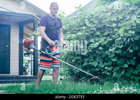 Mann in Brille, Shorts und T-Shirt mäht Rasen mit Benzin Trimmer vor dem Hintergrund des Weinbergs und Eingang zum Haus Stockfoto