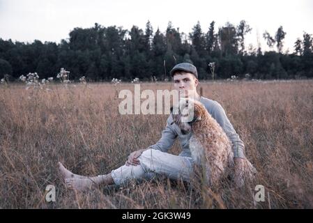 Ein 18-jähriger Junge mit Mütze sitzt an einem Sommerabend mit einem Hund auf einem Feld im dicken Gras. Stockfoto