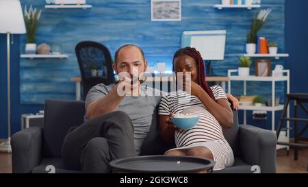 POV von interracial paar mit Schwangerschaft Fernsehen und Popcorn essen. Multiethnische Menschen, die ein Kind erwarten, sitzen auf dem Sofa. Partner, die Spaß haben, während sie die Kamera betrachten Stockfoto