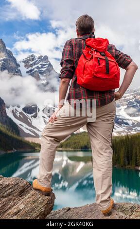 Wandermann mit rotem Rucksack, der auf einem Felsen mit Blick auf den Moraine Lake steht und auf schneebedeckte Rocky Mountain-Gipfel blickt, Banff National Park, A Stockfoto