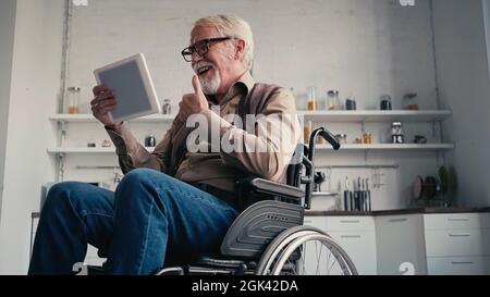 Lächelnder behinderter Rentner im Rollstuhl, der während eines Videoanrufs zu Hause den Daumen hochzeigt Stockfoto