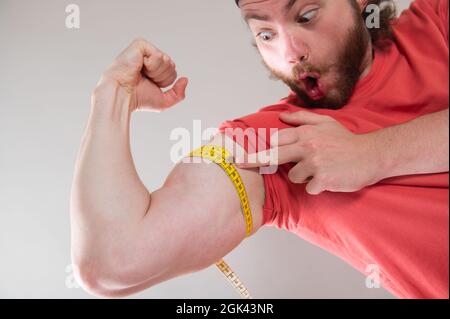 Bärtiger lustiger Mann, der Bizeps, seine Armmuskeln mit einem gelben Maßband misst. Er lächelt, glücklich. Rotes T-Shirt tragen. Hellgrauer Hintergrund. Stockfoto