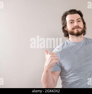 Glücklicher junger bärtiger Mann in T-Shirt gekleidet, zeigt mit Daumen beiseite isoliert auf grauem Studio-Hintergrund Stockfoto