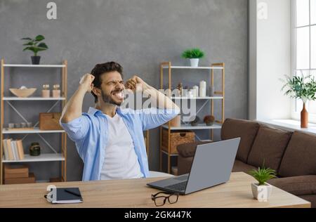 Fröhlicher junger Mann, der mit einem Laptop am Schreibtisch sitzt, sich aufgeregt fühlt, lächelt und mit der Faust pumpt Stockfoto