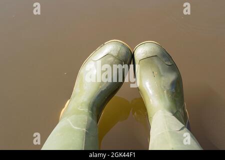 Draufsicht auf ein Paar grüne gummistiefel, die von einer Person über dem Wasser getragen werden. Stockfoto