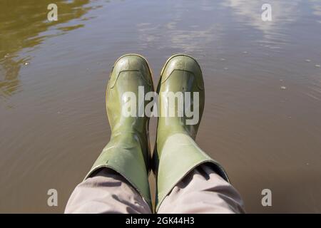 Draufsicht auf ein Paar grüne gummistiefel, die von einer Person über dem Wasser getragen werden. Stockfoto