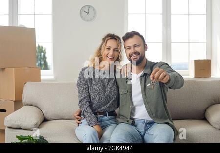 Glückliches junges Paar, das in einer neuen Wohnung auf der Couch sitzt und den Schlüssel zu ihrem neuen Zuhause zeigt Stockfoto