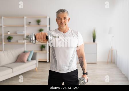 Häusliche Ausbildung mit Gewichten. Positiver reifer Mann, der Übungen mit Hanteln macht und seinen Körper zu Hause stärkt Stockfoto