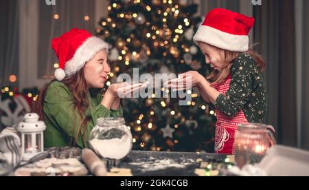 Kleine Mädchen Schwestern in Santa Hüte bläst Mehl auf einander während Prozess Omaking Weihnachtskekse Stockfoto