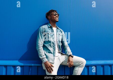 afrikanischer Mann in Sonnenbrille steht gegen eine blaue Wand eines Gebäudes in der Stadt, hübscher schwarzer Mann lächelt Stockfoto