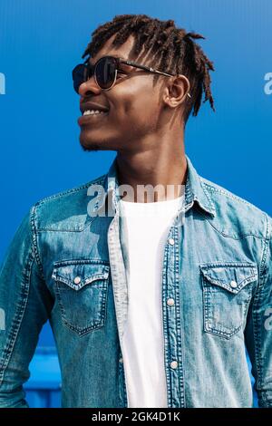 afrikanischer Mann in Sonnenbrille steht gegen eine blaue Wand eines Gebäudes in der Stadt, hübscher schwarzer Mann lächelt Stockfoto