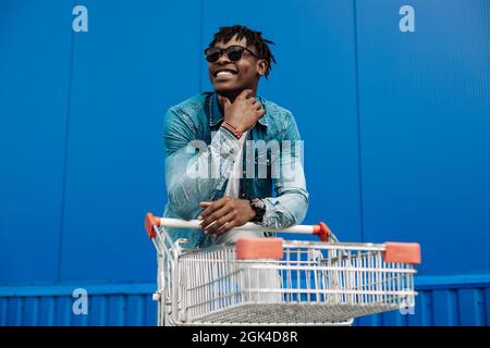 afro-Shopping mit einem Einkaufswagen, der Typ ist in der Nähe des Einkaufszentrums, ein junger schwarzer Student auf dem Hintergrund eines blauen Gebäudes Stockfoto