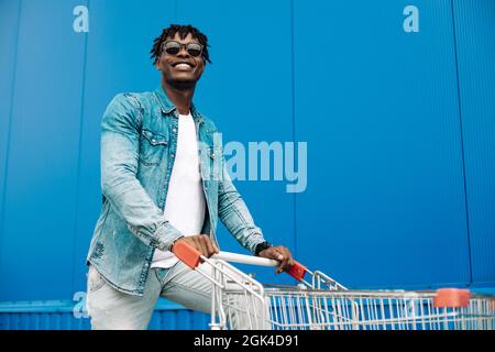 afro-Shopping mit einem Einkaufswagen, der Typ ist in der Nähe des Einkaufszentrums, ein junger schwarzer Student auf dem Hintergrund eines blauen Gebäudes Stockfoto