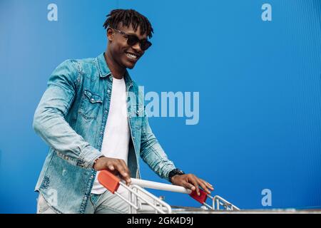 afro-Shopping mit einem Einkaufswagen, der Typ ist in der Nähe des Einkaufszentrums, ein junger schwarzer Student auf dem Hintergrund eines blauen Gebäudes Stockfoto