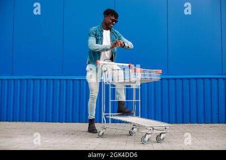 afro-Shopping mit einem Einkaufswagen, der Typ kauft in der Nähe des Einkaufszentrums ein, schaut auf die Uhr an seiner Hand, Konzept: Ladeneröffnung Stockfoto