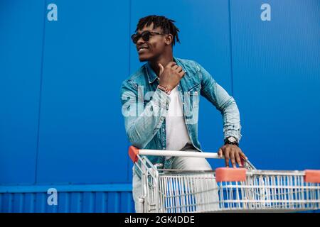 afro-Shopping mit einem Einkaufswagen, der Typ ist in der Nähe des Einkaufszentrums, ein junger schwarzer Student auf dem Hintergrund eines blauen Gebäudes Stockfoto