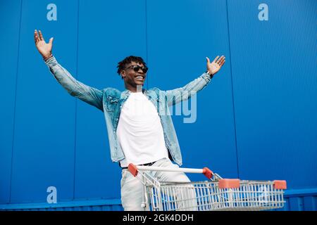 afro-Shopping mit einem Einkaufswagen, der Typ ist in der Nähe des Einkaufszentrums einkaufen, freut sich und hebt die Hände hoch, Konzept: Erfolgreicher Einkauf Stockfoto