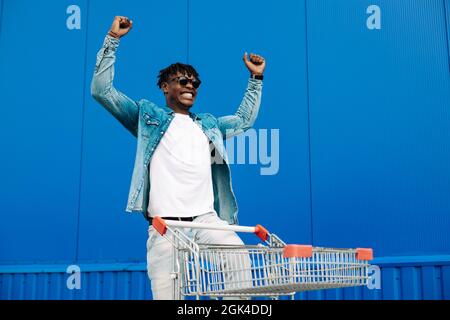 afro-Shopping mit einem Einkaufswagen, der Typ ist in der Nähe des Einkaufszentrums einkaufen, freut sich und hebt die Hände hoch, Konzept: Erfolgreicher Einkauf Stockfoto