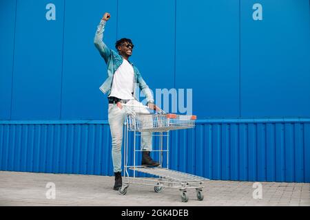 afro-Shopping mit einem Einkaufswagen, der Typ ist in der Nähe des Einkaufszentrums einkaufen, freut sich und hebt die Hände hoch, Konzept: Erfolgreicher Einkauf Stockfoto