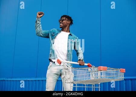 afro-Shopping mit einem Einkaufswagen, der Typ ist in der Nähe des Einkaufszentrums einkaufen, freut sich und hebt die Hände hoch, Konzept: Erfolgreicher Einkauf Stockfoto