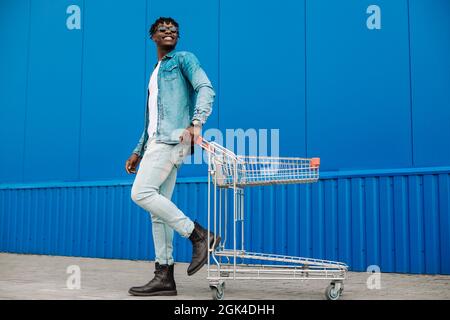 Schwarzer Afro-Mann, der mit einem Einkaufswagen kauft, posiert, Mode, in der Nähe des Einkaufszentrums einkaufen, junger schwarzer Student auf dem Hintergrund eines blauen Bui Stockfoto