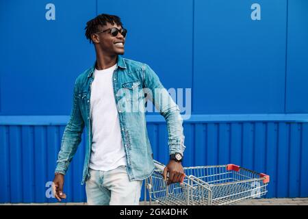 Schwarzer Afro-Mann, der mit einem Einkaufswagen kauft, posiert, Mode, in der Nähe des Einkaufszentrums einkaufen, junger schwarzer Student auf dem Hintergrund eines blauen Bui Stockfoto