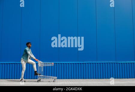 Schwarzer Afro-Kerl läuft mit einem Einkaufswagen einkaufen, der Kerl hat Spaß und springt, läuft für Einkäufe in der Nähe der Mall, junge schwarze Student auf dem Hintergrund eines Stockfoto