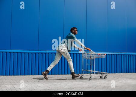 Schwarzer Afro-Kerl läuft mit einem Einkaufswagen einkaufen, der Kerl hat Spaß und springt, läuft für Einkäufe in der Nähe der Mall, junge schwarze Student auf dem Hintergrund eines Stockfoto