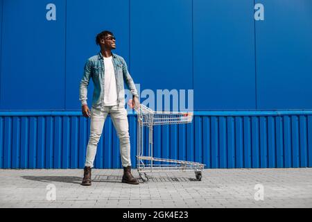 Schwarzer Afro-Mann, der mit einem Einkaufswagen kauft, posiert, Mode, in der Nähe des Einkaufszentrums einkaufen, junger schwarzer Student auf dem Hintergrund eines blauen Bui Stockfoto