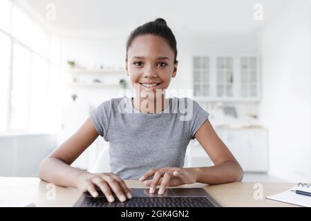 Lächelndes, jugendliches afroamerikanisches Mädchen, das auf der Tastatur tippt und am Tisch mit dem Laptop studiert Stockfoto