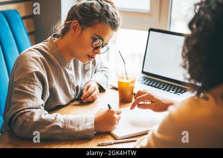 Junge kaukasische Frau und Mann arbeiten und studieren im Café mit Laptop, diskutieren Aufgaben und Vorbereitung auf die Prüfung. Online-Schulungen und Internetkurse Stockfoto