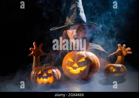 Eine ominöse Hexe im Hut zaubert über eine Jack-o-Laterne. Traditionelle halloween-Figuren. Mystischer Nebel kriecht über Kürbisse mit geschnitzten schrecklich Stockfoto