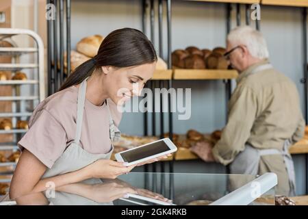 Lächelnde junge Bäckerin auf der Schürze, die mit einem Tisch gegen ihren reifen männlichen Kollegen auflegte, der frisches Brot auf das Regal stellte Stockfoto