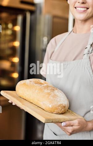 Nahaufnahme einer lächelnden Bäckerin in der Schürze, die weißen Laib Brot auf Holzbrett hielt Stockfoto