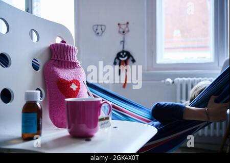 Berlin, Deutschland. September 2021. Ein Fieberthermometer, Hustensirup, eine Heißwasserflasche und eine Tasse Tee stehen auf einem Stuhl, während ein Kind in einer Hängematte im Hintergrund liegt. Quelle: Annette Riedl/dpa/Alamy Live News Stockfoto