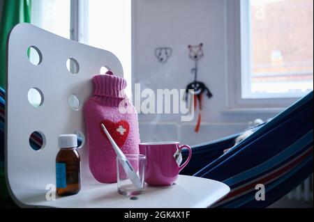 Berlin, Deutschland. September 2021. Ein Fieberthermometer, Hustensirup, eine Heißwasserflasche und eine Tasse Tee stehen auf einem Stuhl, während ein Kind in einer Hängematte im Hintergrund liegt. Quelle: Annette Riedl/dpa/Alamy Live News Stockfoto