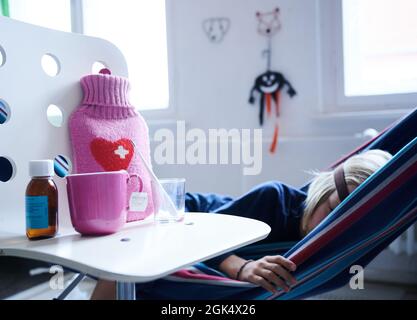 Berlin, Deutschland. September 2021. Ein Fieberthermometer, Hustensirup, eine Heißwasserflasche und eine Tasse Tee stehen auf einem Stuhl, während ein Kind in einer Hängematte im Hintergrund liegt. Quelle: Annette Riedl/dpa/Alamy Live News Stockfoto