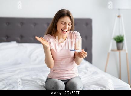 Emotionale tausendjährige Dame überprüft Schwangerschaftstest, glücklich über zukünftige Mutterschaft zu Hause Stockfoto