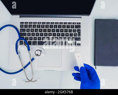 Hand mit blauem Nitrilhandschuh, der einen gebrauchten Antigentest hält. Stethoskop für Laptop und Notebook. Stockfoto