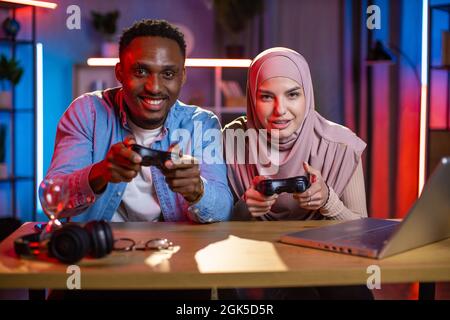 Junges multirassisches Paar, das mit drahtlosen Joysticks in den Händen am Tisch sitzt und Videospiele auf dem Laptop spielt. Afrikanischer Mann und muslimische Frau verbringen abends viel Spaß zu Hause. Stockfoto