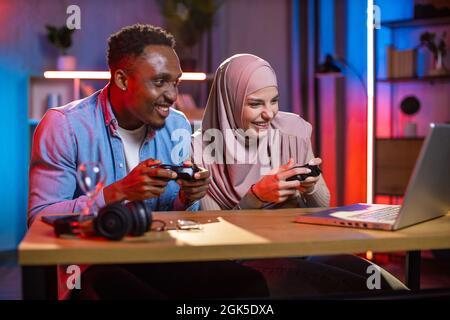 Aufgeregt multiethnischen Paar mit Joysticks in den Händen Blick auf den Computerbildschirm, während Videospiele spielen. Konkurrenzmodus des afrikanischen Mannes und der muslimischen Frau. Häusliches Leben mit Spaß. Stockfoto