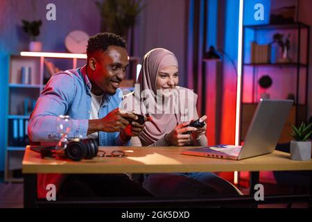 Aufgeregt multiethnischen Paar mit Joysticks in den Händen Blick auf den Computerbildschirm, während Videospiele spielen. Konkurrenzmodus des afrikanischen Mannes und der muslimischen Frau. Häusliches Leben mit Spaß. Stockfoto
