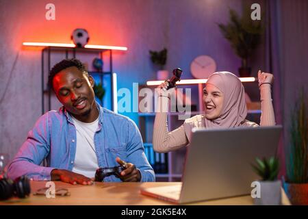 Glückliches multiethnisches Paar, das am Abend einen kabellosen Laptop und Joysticks zum Spielen von Videospielen verwendet. Muslimische Frau im Hijab gewinnt im Wettbewerb mit afrikanischem Mann. Stockfoto