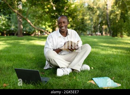 Glücklicher afroamerikanischer Kerl, der mit Buch und Laptop im Park studiert, auf dem College-Campus sitzt und sich Notizen macht Stockfoto