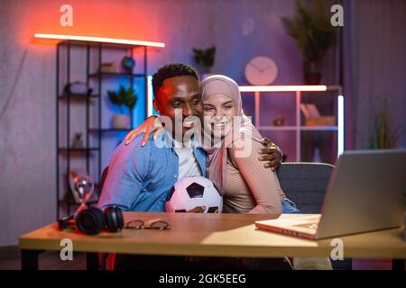 Positives multikulturelles Paar, das in Umarmungen am Tisch sitzt, während es sich ein Fußballspiel auf einem kabellosen Laptop ansieht. Unterhaltung am Abend zu Hause. Fußball-Fan-Konzept. Stockfoto