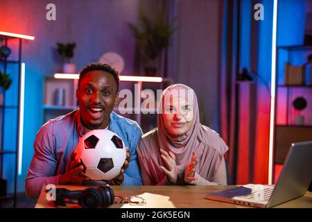 Porträt eines afrikanischen Mannes und einer muslimischen Frau, die während des Fußballspiels ihre Lieblingsmannschaft emotional jubelten. Junges Paar mit einem modernen Laptop, um sich ein Spiel zu Hause anzusehen. Stockfoto