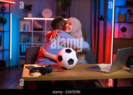Glückliche arabische Frau in Hijab umarmt sich mit afroamerikanischem Mann, während sie Fußballspiele auf dem Laptop ansieht. Junges Paar feiert den Sieg der Lieblingsmannschaft. Konzept für Unterstützung und Emotionen. Stockfoto