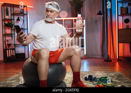 Grauhaariger kaukasischer Mann in Sportkleidung sitzt auf einem Fitball mit einer Flasche Wasser in der Hand. Gealterter Mann, der Selfie auf einem modernen Smartphone nimmt, während er sich nach dem Training entspannt. Stockfoto