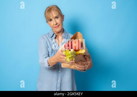 Ältere Frau, die Lebensmittel einkaufen kann. Isoliert auf blauem Hintergrund. Stockfoto